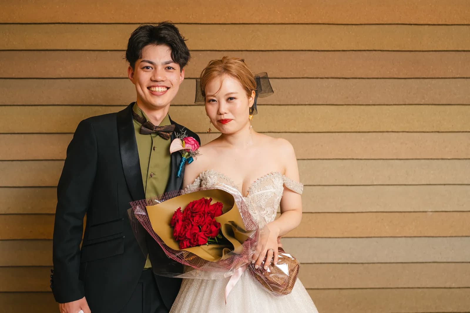 木目調の壁の前で赤いバラの花束を持つ新郎新婦