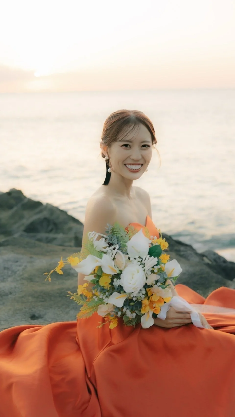 オレンジのドレスを着た花嫁が夕景の海辺でブーケを持つ様子
